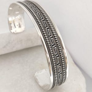 May include: A silver cuff bracelet featuring a central Greek key pattern flanked by two twisted rope-style borders. The metal has a polished finish with darkened recessed areas to highlight the intricate geometric and linear detailing.
