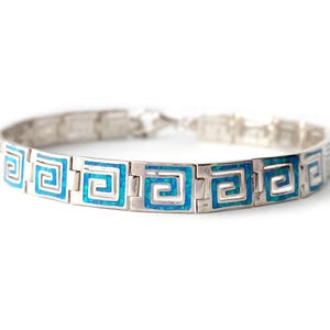 Pode incluir: Pulseira prateada com um design de chave grega repetido com incrustações azuis vibrantes. A pulseira é composta por elos retangulares, cada um com o padrão geométrico. O fecho é visível na extremidade da pulseira.