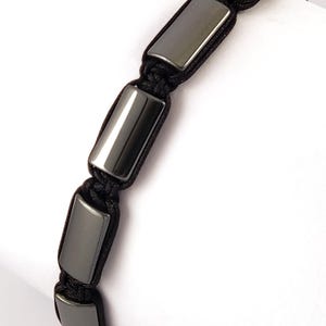 Peut inclure: Un bracelet noir composé de perles rectangulaires en hématite polie gris foncé. Les perles sont enfilées sur un cordon noir, créant un design minimaliste et moderne. Le bracelet est présenté sur fond blanc.