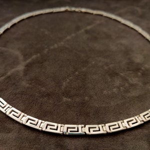 May include: A silver necklace with a Greek key pattern. The necklace is on a brown fabric background.