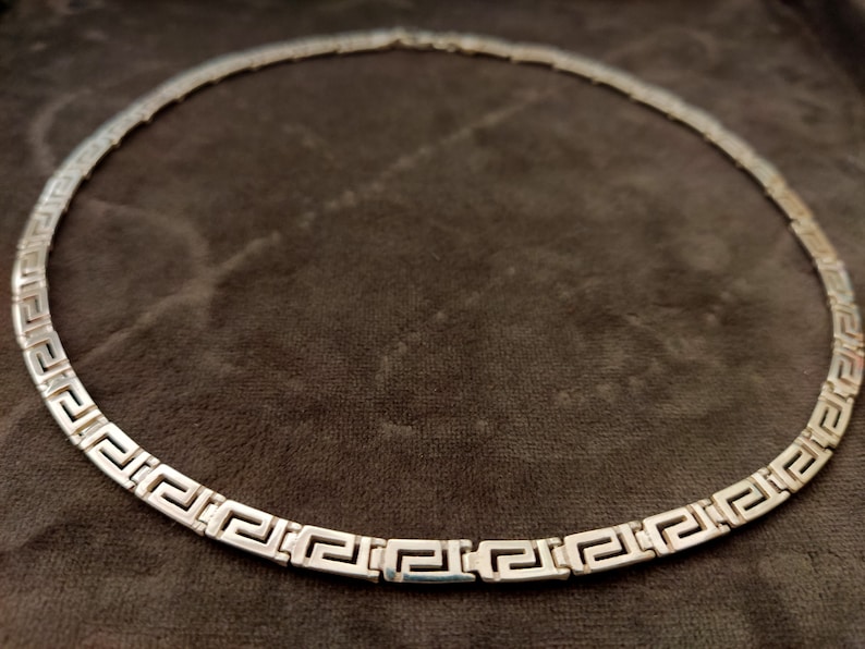 May include: A silver necklace with a Greek key pattern. The necklace is on a brown fabric background.