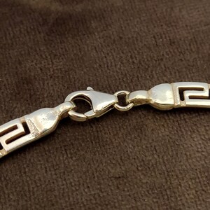 May include: Silver chain necklace with a Greek key pattern and a lobster clasp closure.