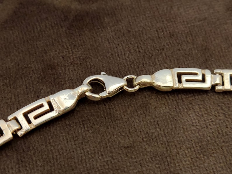 May include: Silver chain necklace with a Greek key pattern and a lobster clasp closure.