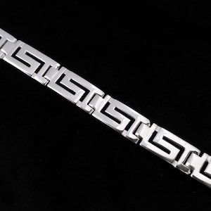 May include: A silver-toned bracelet with a Greek key design. The bracelet is made of metal and features a repeating geometric pattern. The background is black, highlighting the bracelet's bright, reflective surface. This accessory is suitable for various occasions.