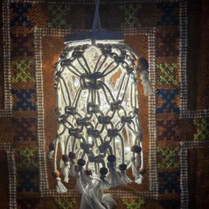 May include: A handmade macrame jar lamp with a warm glow. The white macrame design covers a clear glass jar, adorned with wooden beads and tassels. The lamp hangs from a white cord, creating a cozy, bohemian aesthetic.
