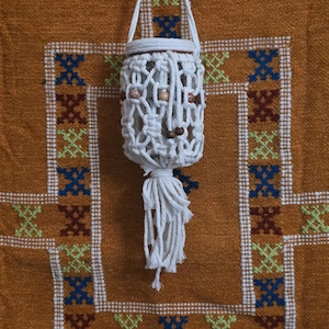 Pode incluir: Um suporte de plantas em macramé branco com contas de madeira e uma borla, suspenso contra um fundo laranja texturizado. O suporte de plantas foi projetado para conter um pequeno vaso ou recipiente. O fundo apresenta um padrão geométrico.