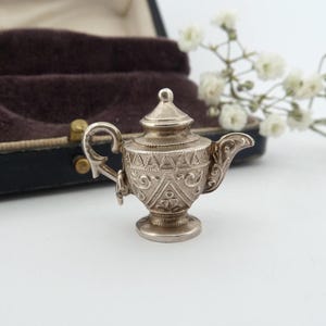 May include: A miniature silver-toned teapot charm featuring an ornate, embossed pattern of triangles and swirling filigree details. The small vessel has a pointed lid, a curved handle, and a decorative spout, showcasing a metallic finish.