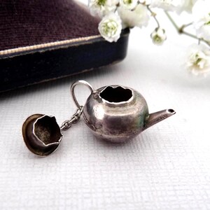 May include: A miniature silver-toned metal teapot charm featuring a rounded body, a small spout, and a removable lid attached by a delicate metal chain. The teapot has a tarnished metallic finish with visible signs of age and oxidation.
