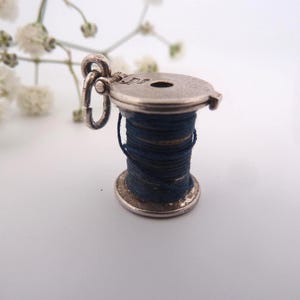 May include: A vintage silver-toned charm in the shape of a thread spool. The spool is filled with dark blue thread and has a small loop for attachment. Delicate white flowers are in the background.