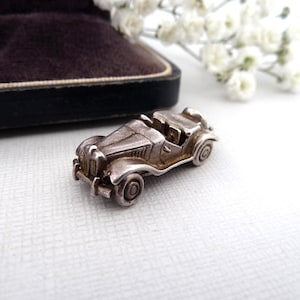 May include: A small, silver-toned metal charm shaped like a vintage open-top roadster car. The miniature vehicle features detailed fenders, a front grille, circular wheels, and an open cockpit design with a visible steering wheel and seats.