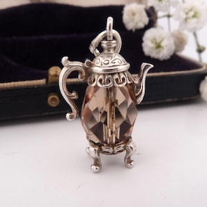 May include: A silver teapot charm with a faceted, smoky quartz-coloured body. The charm has ornate detailing on the handle, spout, and lid, and sits on three small feet. A loop is at the top for attachment.