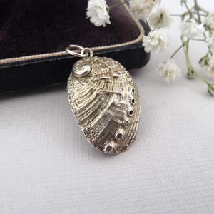 May include: A silver abalone shell pendant. The pendant has a textured surface and small holes. It hangs from a silver loop. The background includes a dark velvet box and white flowers. The pendant is a unique piece of jewellery.