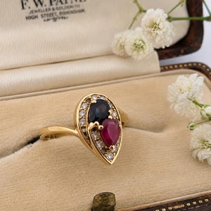 May include: A gold ring featuring a pear-shaped dark blue sapphire and a pear-shaped red ruby, surrounded by a halo of small round white diamonds. The ring is set in a yellow gold band and displayed inside a vintage jewelry box with cream lining.