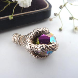 May include: A silver-toned claw grasping a faceted, iridescent crystal. The crystal reflects shades of purple, blue, and green. The claw has detailed texture, and the background is a soft white with hints of purple and white flowers.