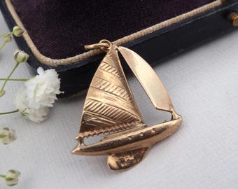 May include: A gold-toned metal sailboat pendant featuring a textured triangular sail and a smooth hull. The pendant includes a small jump ring at the top for attachment. The design depicts a stylized vessel with a single mast and two distinct sails.