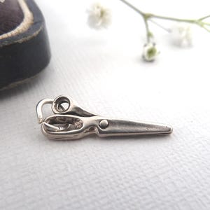 Könnte beinhalten: Ein kleiner, silberfarbener Anhänger in Form einer Schere. Der Anhänger hat oben eine Schlaufe zum Befestigen an einer Kette oder einem Armband. Die Schere ist mit einem kleinen Niet und einem gebogenen Griff versehen. Ein Schmuckstück.