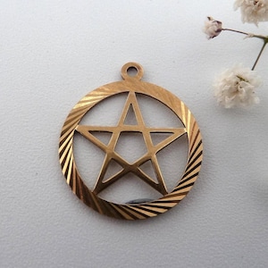 May include: A gold-toned metal pendant featuring a five-pointed star enclosed within a circular frame. The circular border displays a radial, sunburst-like etched pattern, and a small circular loop is attached at the top for hanging purposes.