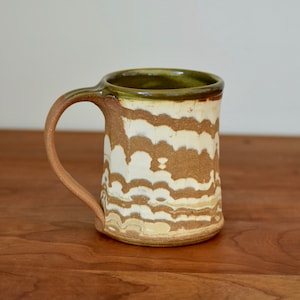 May include: A brown and white ceramic mug with a green glaze on the rim. The mug has a unique, wavy pattern.