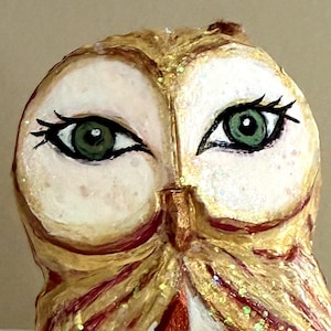 May include: A hand-painted owl figurine featuring a gold and tan textured finish with glitter accents. The owl has large, detailed green eyes with black eyelashes and a chest area decorated with a white and copper-colored checkered pattern.