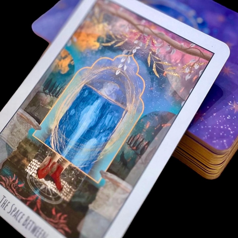 May include: A tarot card featuring a blue portal with a waterfall design, framed by a gold archway. The card has a white border and the text "THE SPACE BETWEEN". A deck of cards with gold edges is visible.