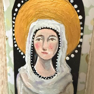 May include: A painted religious icon featuring a woman with a gold halo and a white veil. The woman has a serene expression, with a gold cross on her chest. The background is black with white dots, and the overall style is folk art.