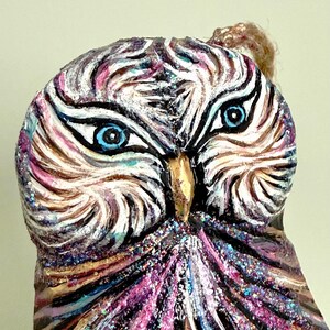 May include: A hand-painted owl ornament featuring vibrant purple, pink, and white brushstrokes with glitter accents. The owl has piercing bright blue eyes, a gold-toned beak, and a textured surface. A small jute twine loop is attached at the top.