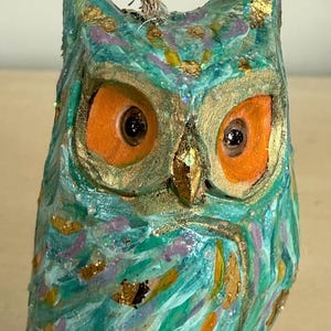 May include: A hand-painted owl ornament featuring a textured teal and turquoise surface with accents of purple, gold leaf flakes, and glitter. The owl has large, bright orange eyes with dark pupils and a gold-painted beak, suspended by a jute twine loop.