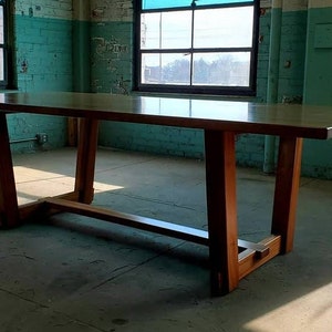 May include: A large, rectangular wooden table with a light brown top and a unique, modern design. The table has a simple, geometric base with four legs that are connected by a single, horizontal beam. The table is shown in a room with a concrete floor and green walls.