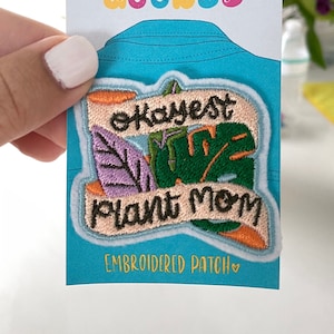 May include: A pink and orange embroidered patch with green and purple leaves and the text "Okayest Plant Mom" with the words "Embroidered Patch" below.