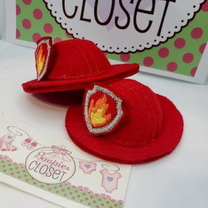 May include: Two small red felt firefighter hats with embroidered flame patches. The hats are displayed on a white surface with a sign that says "Boopies Closet" and a sign that says "closet".