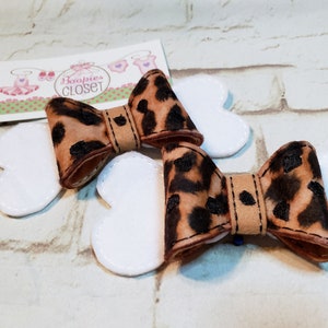 May include: Two brown and black leopard print hair bows with white felt backing. The bows are attached to a white felt bone shape.