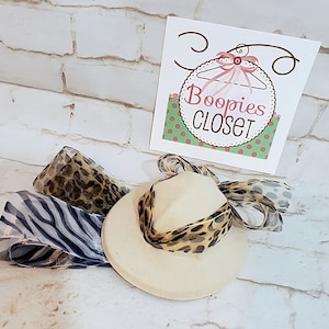 May include: A white fedora hat with a leopard print ribbon tied around the crown. The hat is sitting on a white brick background with a sign that says "Boopies Closet".