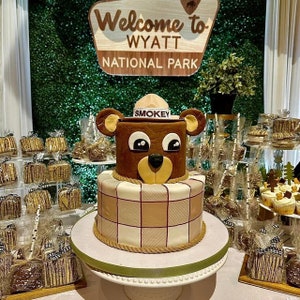 May include: A two-tiered cake decorated with a brown bear wearing a ranger hat with the word "SMOKEY" on it. The bottom tier is covered in a plaid pattern. The cake is on a white cake stand with a green ribbon. The sign behind the cake says "Welcome to WYATT NATIONAL PARK".