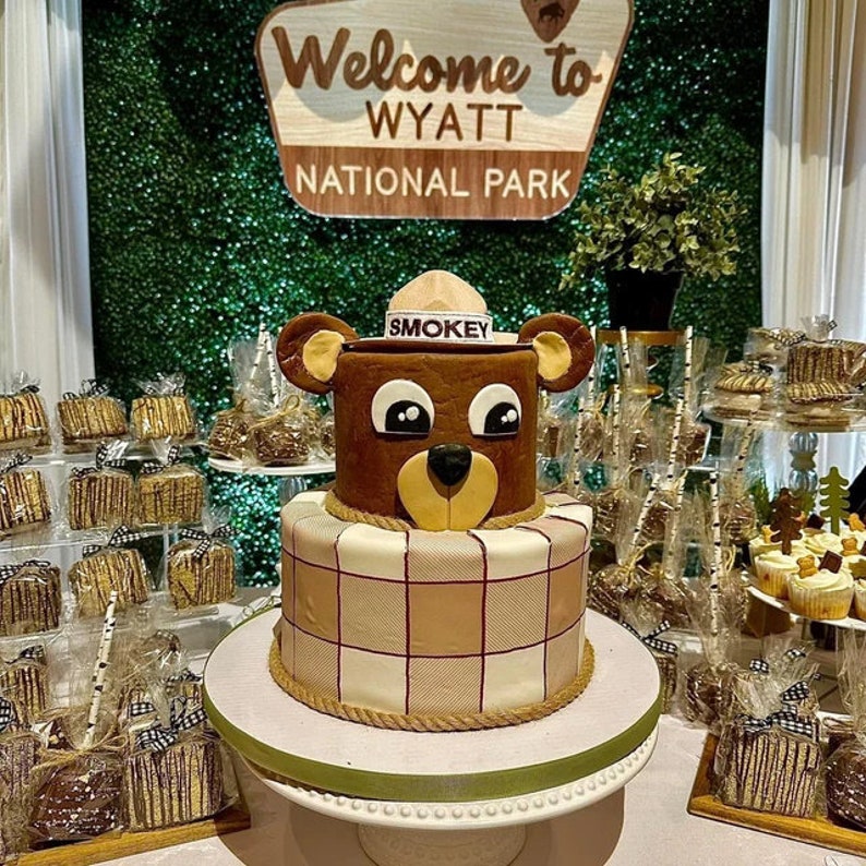 May include: A two-tiered cake decorated with a brown bear wearing a ranger hat with the word "SMOKEY" on it. The bottom tier is covered in a plaid pattern. The cake is on a white cake stand with a green ribbon. The sign behind the cake says "Welcome to WYATT NATIONAL PARK".