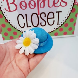 May include: A small blue hat with a white daisy on top. The hat has a black ribbon around the base. The hat is sitting on a hand.