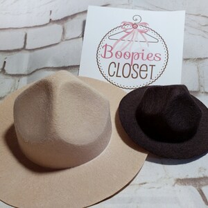 May include: Two felt hats, one beige and one brown, with a wide brim and a pointed crown. The hats are on a white brick background.