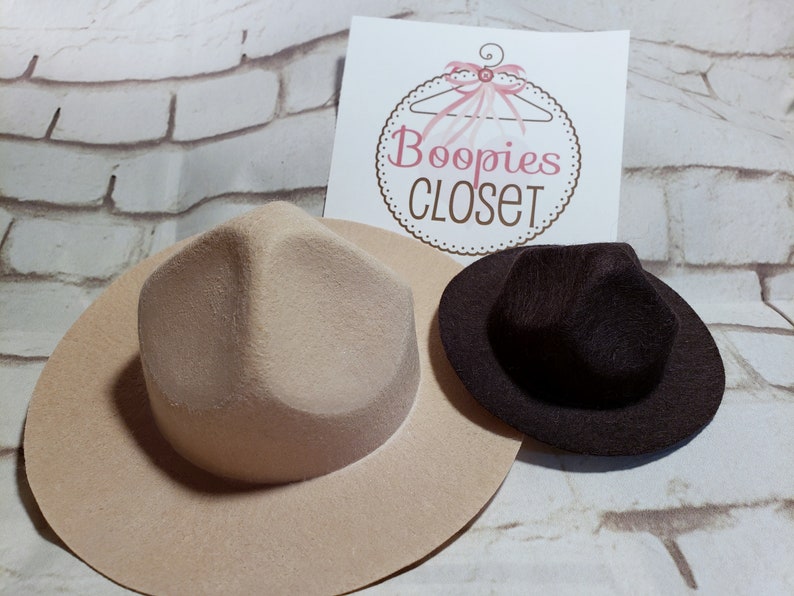 May include: Two felt hats, one beige and one brown, with a wide brim and a pointed crown. The hats are on a white brick background.