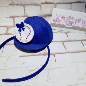 May include: Blue felt baseball cap headband with a white baseball and red stitching. The headband is blue and the cap has a small blue bow on the side.