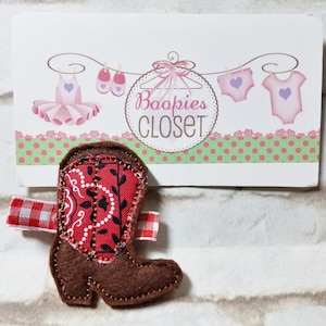 May include: A brown felt cowboy boot with a red and white bandana pattern on the shaft. The boot is a hair clip and has a red and white gingham ribbon attached to the back.