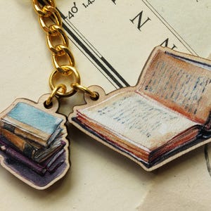 Könnte beinhalten: Eine goldfarbene Kette hält zwei Holzanhänger in Buchform. Ein Anhänger zeigt einen Stapel Bücher mit blauen, braunen und violetten Einbänden. Der andere Anhänger zeigt ein offenes Buch mit detaillierten Seiten. Der Hintergrund ist eine Vintage-Karte.