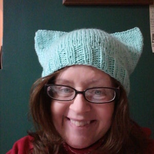 Puede incluir: Un gorro de punto azul turquesa con orejas de gato. El gorro está hecho de un hilo suave y texturizado y tiene un diseño simple y clásico.