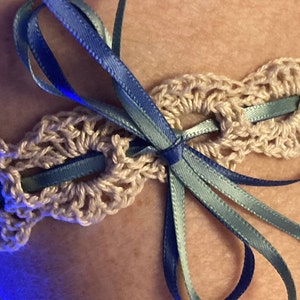 May include: A crocheted garter with a blue and teal ribbon tied in a bow. The garter is made of beige yarn and has a delicate, lacy design.