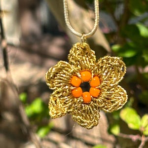 May include: A gold chain necklace with a crocheted flower pendant. The flower is made of gold thread and has a center of orange beads.
