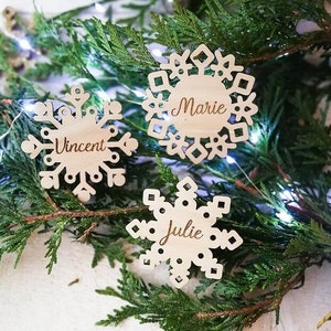Peut inclure: Trois décorations en forme de flocons de neige en bois, gravées des noms "Vincent", "Marie" et "Julie". Les flocons de neige brun clair sont posés sur des branches de pin vertes et un mur de briques blanches.