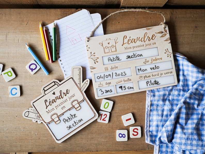 May include: Wooden sign with a school-themed design, featuring a backpack, ruler, and pencil. The sign has blank spaces for writing the child's name, date, age, height, and favorite things. The text on the sign reads "L&eacute;andre, Mon premier jour en Petite section, j'adore Mon velo, plus tard je serai Pilote".