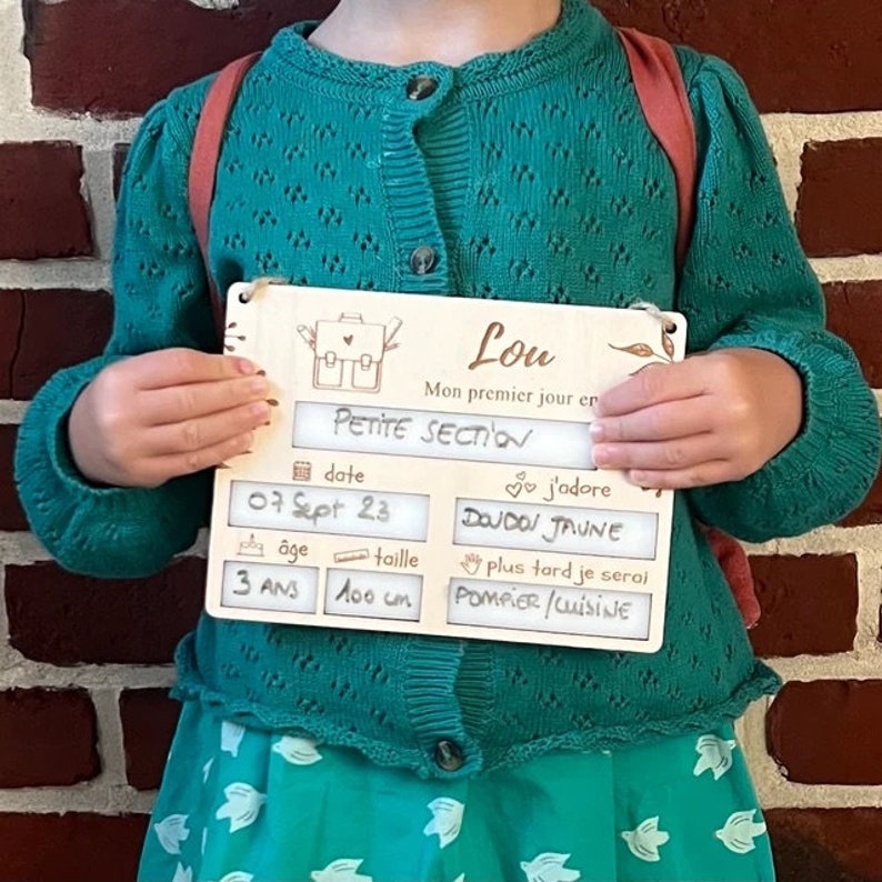 May include: A wooden sign with a light brown background and a white border. The sign has a picture of a backpack with a heart on it. The sign says "Lou" and "Mon premier jour en Petite Section". There are also sections for the date, age, and height. The date is filled in with "07 Sept 23". The age is filled in with "3 ans" and the height is filled in with "100 cm". There is a section for "j'adore" and it is filled in with "DOUDOU JAUNE". There is a section for "plus tard je serai" and it is filled in with "POMPIER/CUISINE".