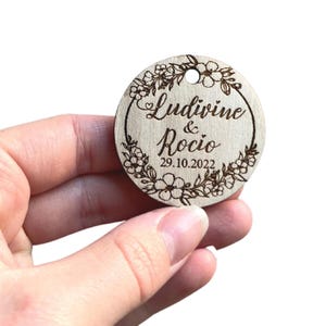 Peut inclure: Une étiquette ronde en bois avec un motif floral et le texte "Ludivine & Rocío 29.10.2022" gravé dessus.