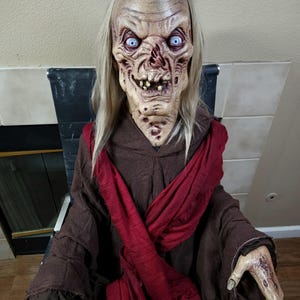 Puede incluir: Una grotesca decoración de Halloween con una cara pálida y arrugada, ojos azules penetrantes y pelo largo y rubio. La figura lleva una túnica marrón con una bufanda roja y tiene manos desfiguradas. Un espeluznante accesorio para Halloween.