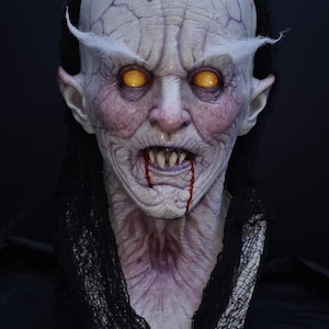 May include: A detailed mask with a pale, grotesque face, yellow eyes, and blood dripping from the mouth. The mask has a wrinkled forehead and is framed by a black hood. Ideal for Halloween or horror events. The mask has sharp teeth.
