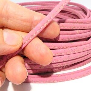 Puede incluir: Cordón de corcho rosa con un detalle de costura dorada. El cordón está enrollado y se sostiene en una mano.